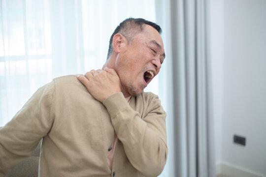 Senior Asian man sitting on chair with hand on neck, expressing neck pain or muscle stiffness. Concept of elderly health, joint problem or physical discomfort at home.