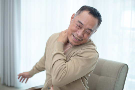 Senior Asian man sitting on chair with hand on neck, expressing neck pain or muscle stiffness. Concept of elderly health, joint problem or physical discomfort at home. - Powered by Adobe