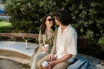 A woman smiles and places her hand on the leg of a man sitting next to her on a bench while they look at each other and hold cups