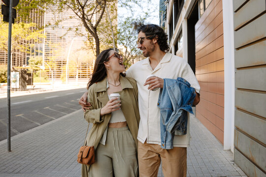 A man hugs a woman walking next to him while they laugh and hold cups