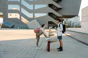 A female athlete ties her shoelaces and rests her foot on a stone stool while laughing and talking to a male athlete standing across from her holding a bottle