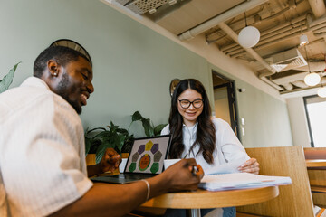 A male worker reads a notebook and sits at a desk next to a female worker who reads documents while...