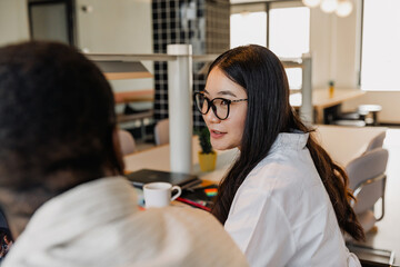 A female employee is smiling and talking to a male employee sitting next to her at the desk