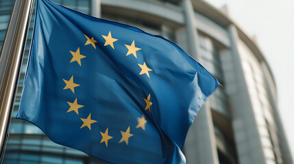 The European flag waives against a modern building. The flag is blue with yellow stars. The building behind it is gray. The flag is on a metal pole and waving in the wind.