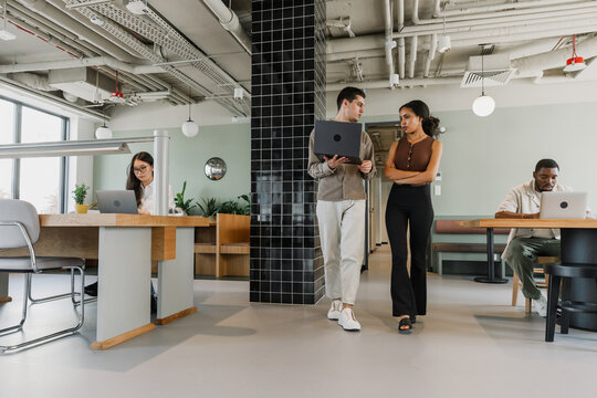 A male worker is holding a laptop while walking next to a female worker while they are talking
