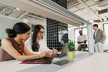 Two female workers talking and sitting at a table while looking at a laptop