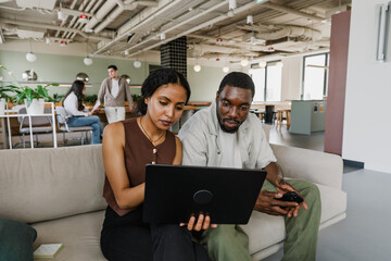 Male and female workers are sitting on the sofa and looking at the laptop that she is showing