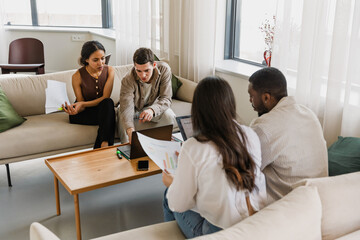 A group of four workers sit on sofas and look at laptops while female workers hold charts