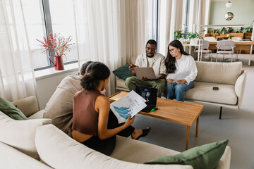 A group of four workers sit on couches while the women hold charts and the man holds a laptop and talks