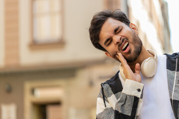 Upset Hispanic adult young man feels a toothache while standing in urban city street. Sad Indian male tourist guy suffering pain holding hand to cheek and massaging. He needs the help of a dentist.