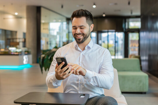 Young smiling Caucasian businessman using smartphone typing text messages, reading email, browsing news in office lobby. Handsome man guy using mobile social media app in hotel hall. Business people.