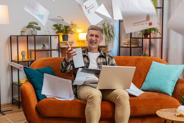 Stressed Caucasian young man freelancer reviewing business charts and graphs and throwing papers unpaid bills in air sitting on couch in home living room. Angry guy remote working on sofa in apartment