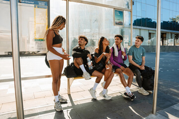 A female athlete stands next to a group of four athletes sitting on a bench at a bus stop while they laugh and talk © Drobot Dean