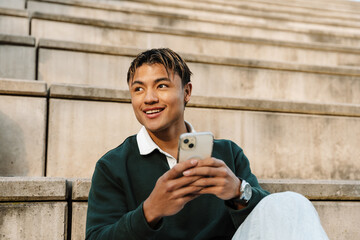 A male student is smiling and sitting on the bleachers while holding the phone
