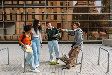 A group of four students stand and laugh while talking and holding laptops and notebooks while two of them lean
