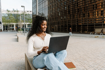 A female student is holding a laptop and listening in earphones while sitting on a bench and smiling