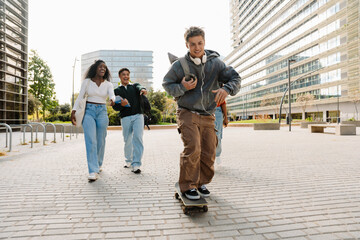 A male student rides a skateboard while a group of three students follow him while they laugh