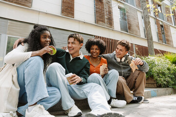 A group of four students laugh and look at one of them while sitting on a curb and holding food