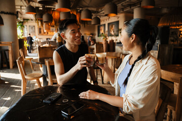 A man and a woman clink cups and laugh while sitting at a table