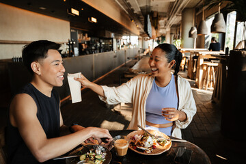 A woman laughs and holds a napkin near the face of a man sitting next to her at the table