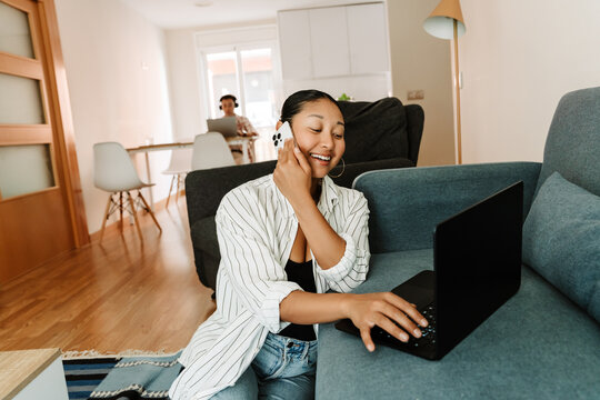 A woman is laughing and talking on the phone while sitting on the carpet and typing on a laptop on the sofa