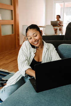 Fototapeta A woman is talking on the phone and smiling while sitting on the floor and typing on a laptop on the sofa