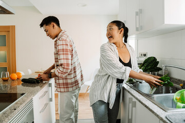 A woman is washing lettuce in the sink and talking to a man who is cutting vegetables behind her
