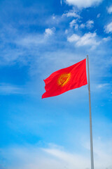 Kyrgyzstan national flag featuring a red background with a traditional tunduk symbol and a yellow sun. Waving prominently on a flagpole against a clear, blue sky. Symbol of unity and heritage