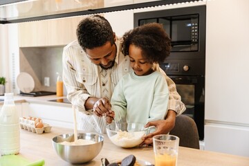 A father hugs his daughter and holds a bowl while he helps her stir in the bowl