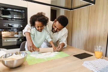 A father helps his daughter roll out dough with a rolling pin while they stand at the table