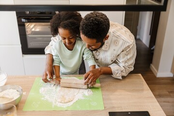 A daughter rolls out dough with a rolling pin while her father helps her, who is standing next to...