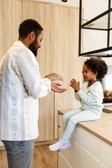 Fototapeta premium A father stands across from his daughter who is sitting on a table while they hold hands facing each other