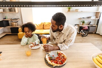 A father is holding a glass and talking to his daughter who is eating while they are sitting at the...