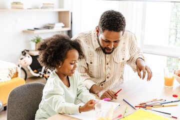 A father is holding a pencil and talking to his daughter who is smiling and holding a pen while...