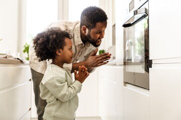 A father and his daughter stand and hold hands together while looking into the oven