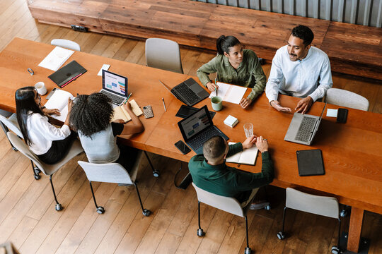 A group of three workers are talking while two female workers are looking at a laptop while sitting at a desk next to them