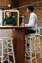 A male employee laughs and listens to a male employee sitting next to him at the table in front of...