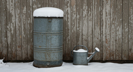 Naklejka premium A vintage metal Snow-covered rain barrel and watering can against a rustic fence, a charming photo for country living and gardening blogs.