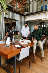 A group of four workers listen to a male worker holding a computer tablet and talking while sitting...