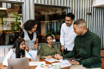 Fototapeta premium A group of five workers are talking and looking at documents on a table with three of them sitting while two of them stand