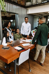 A group of four workers are laughing at a table and talking to a male worker standing across from...