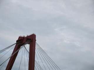 Fototapeta premium Red suspension bridge tower against dramatic cloudy sky, architectural close-up with steel cables. Engineering marvel, iconic structure, urban infrastructure, minimal color contrast.
