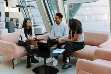 Female worker holding a folder and sitting on a chair while talking to a male and female workers sitting on a couch while he smiles