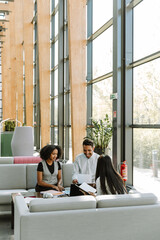 Fototapeta premium Male and female employees laughing and listening to female employee sitting across from them on sofa and holding a notebook