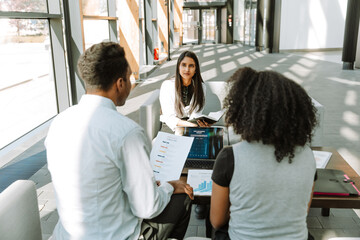 A female worker is holding a notebook and listening to male and female workers sitting across from her on the sofa