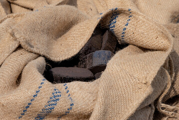 Closeup of dark fuel briquettes partially revealed inside a folded burlap sack, showing coarse jute texture and compacted block shapes