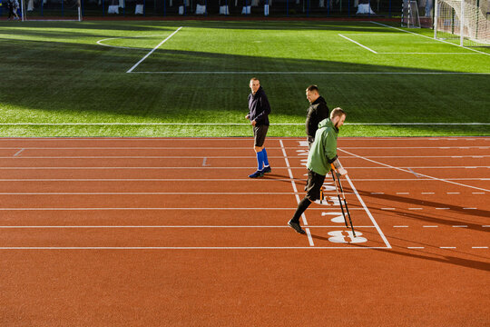 A group of three men walk along a running track while one of them talks and two of them hold crutches - Powered by Adobe