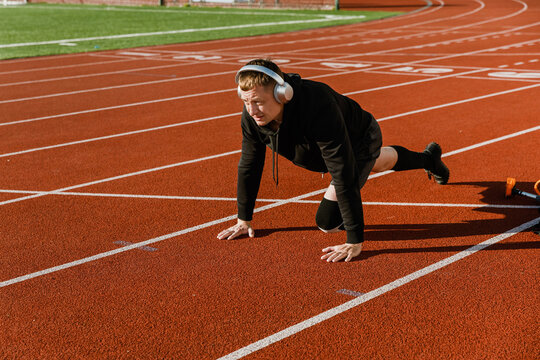 A man rests his hands on the running track and listens in headphones - Powered by Adobe