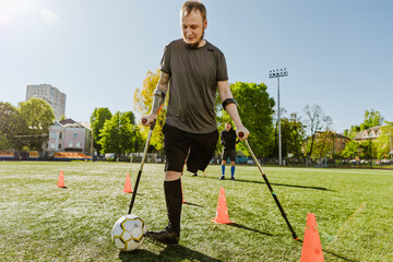 A man dribbles a ball near the cones and leans on the field with crutches
