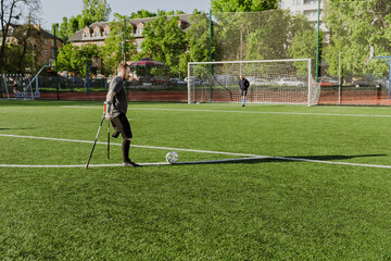 A man on crutches is about to kick a ball into a goal on which a man is standing